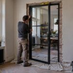Monteur zet een stalen deur met glas in een kale doorgang, waterpas op de deur, afdekzeilen over de visgraatvloer